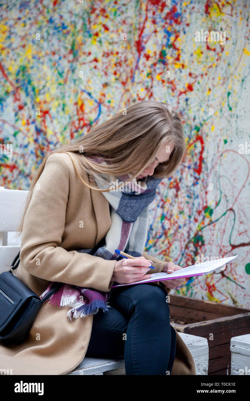 Giovane donna bionda posa sul banco e la scrittura. Donna in cappotto e sciarpa iscritto all'aperto. Foto Stock