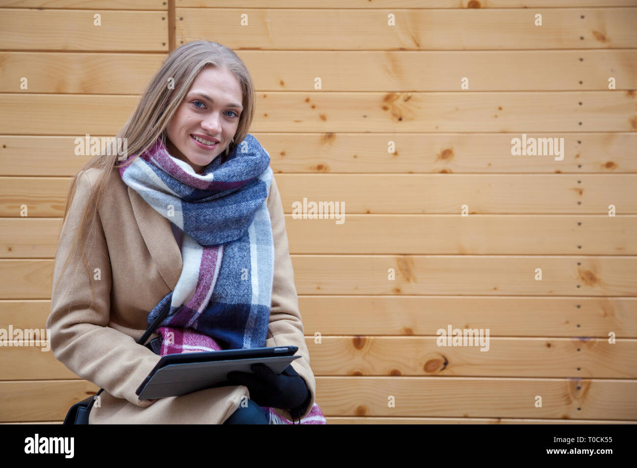 Bionda bella donna azienda tablet all'aperto. Ritratto di giovane donna indossa foulard tartan nella parte anteriore della parete di legno e utilizzando un tablet. Foto Stock