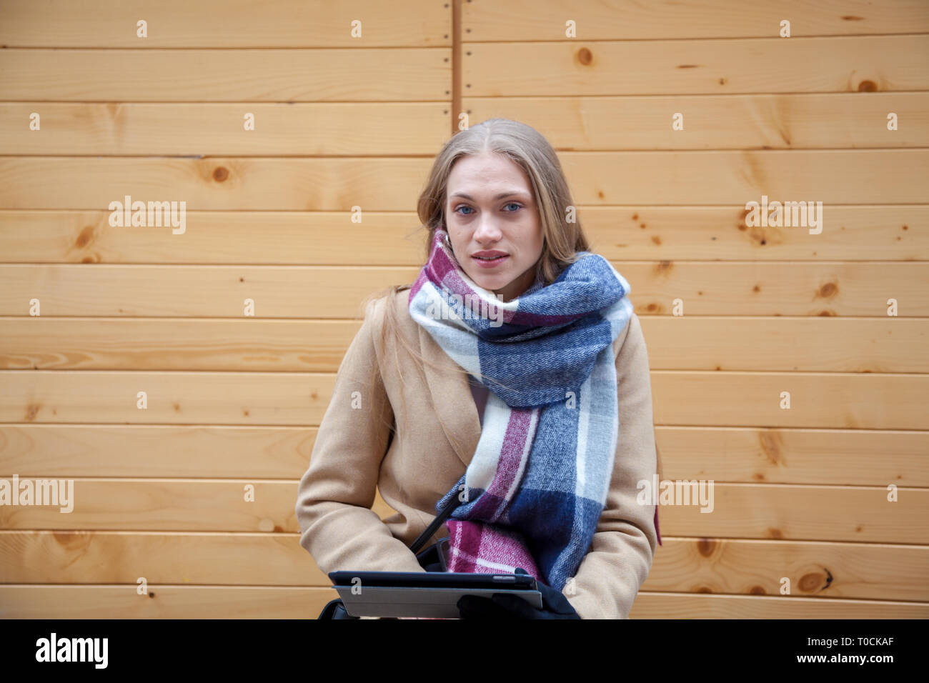 Bionda bella donna azienda tablet all'aperto. Ritratto di giovane donna indossa foulard tartan nella parte anteriore della parete di legno e utilizzando un tablet. Foto Stock