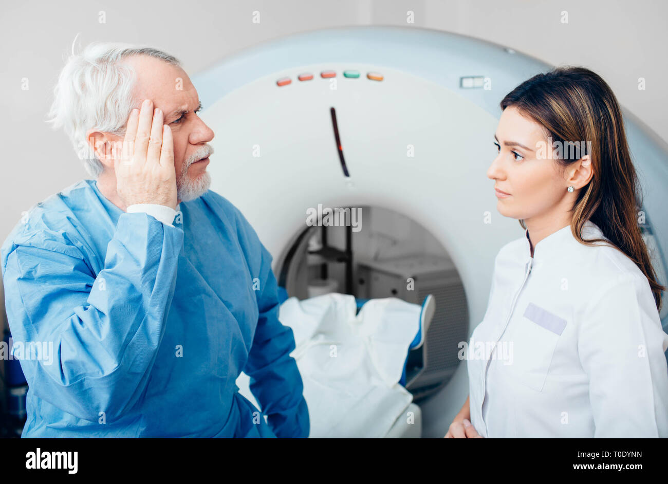 Senior denuncia del paziente di dolore di testa al suo medico mentre è seduto sulla tavola dello scanner. Foto Stock