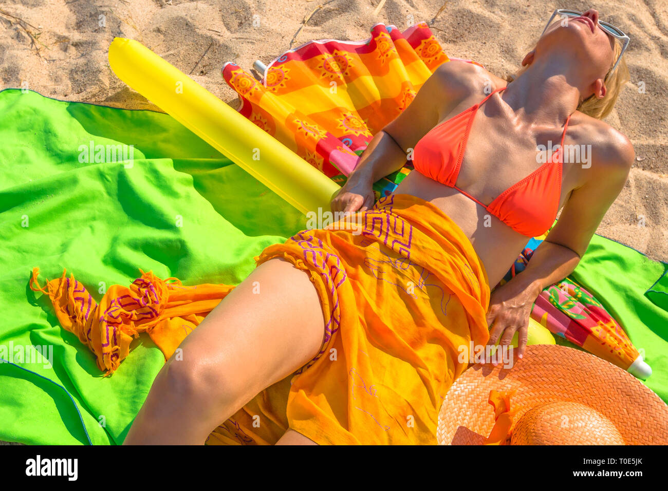 Una donna la concia sdraiato sulla spiaggia con i vestiti e le attrezzature da mare arancio brillante, i colori verde e giallo. Tra gli oggetti: sarong, cappello, costume da bagno Foto Stock