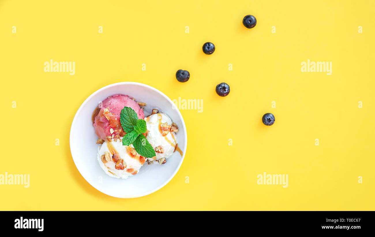 Vanila e gelato alla fragola con caramello e menta su bianco ciotola con dadi e mirtilli su sfondo giallo. Tre convogliatori, splendidi dessert. Scatto dall'alto. Foto Stock