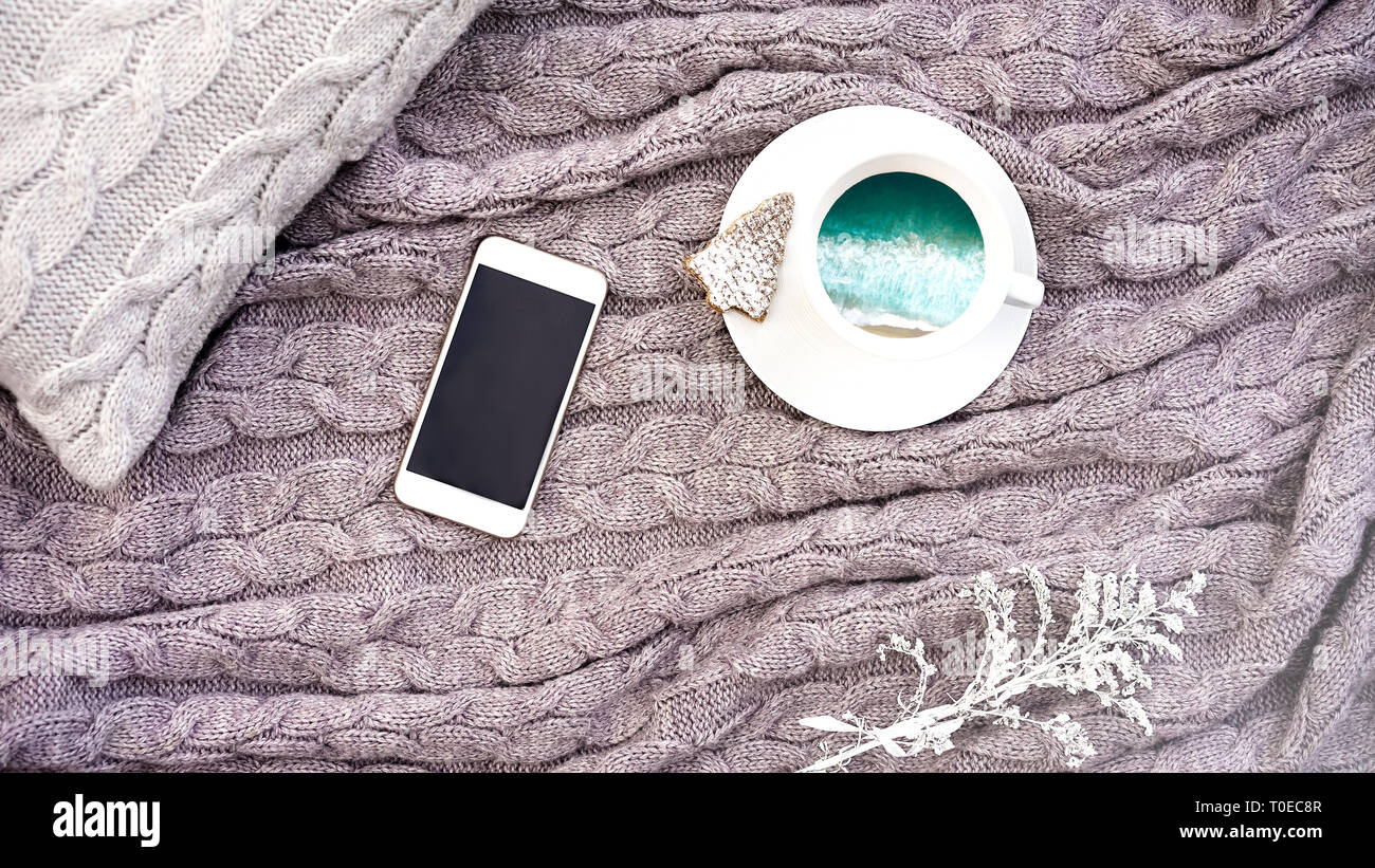 White tazza di caffè con mare turchese onde e waffle cookie nella forma di abete bianco e fiore telefono sul cuscino e caldo grigio di lana plaid. Tessile lavorato a maglia con motivo a treccia. Autunno Inverno concetto comfort Foto Stock