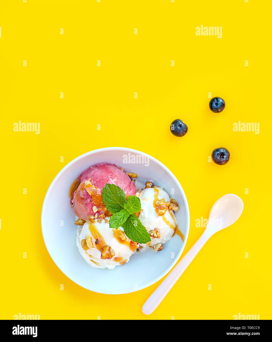 Vanila e gelato alla fragola con caramello e menta su bianco ciotola con dadi e mirtilli su sfondo giallo. Tre convogliatori, splendidi dessert. Scatto dall'alto. Foto Stock