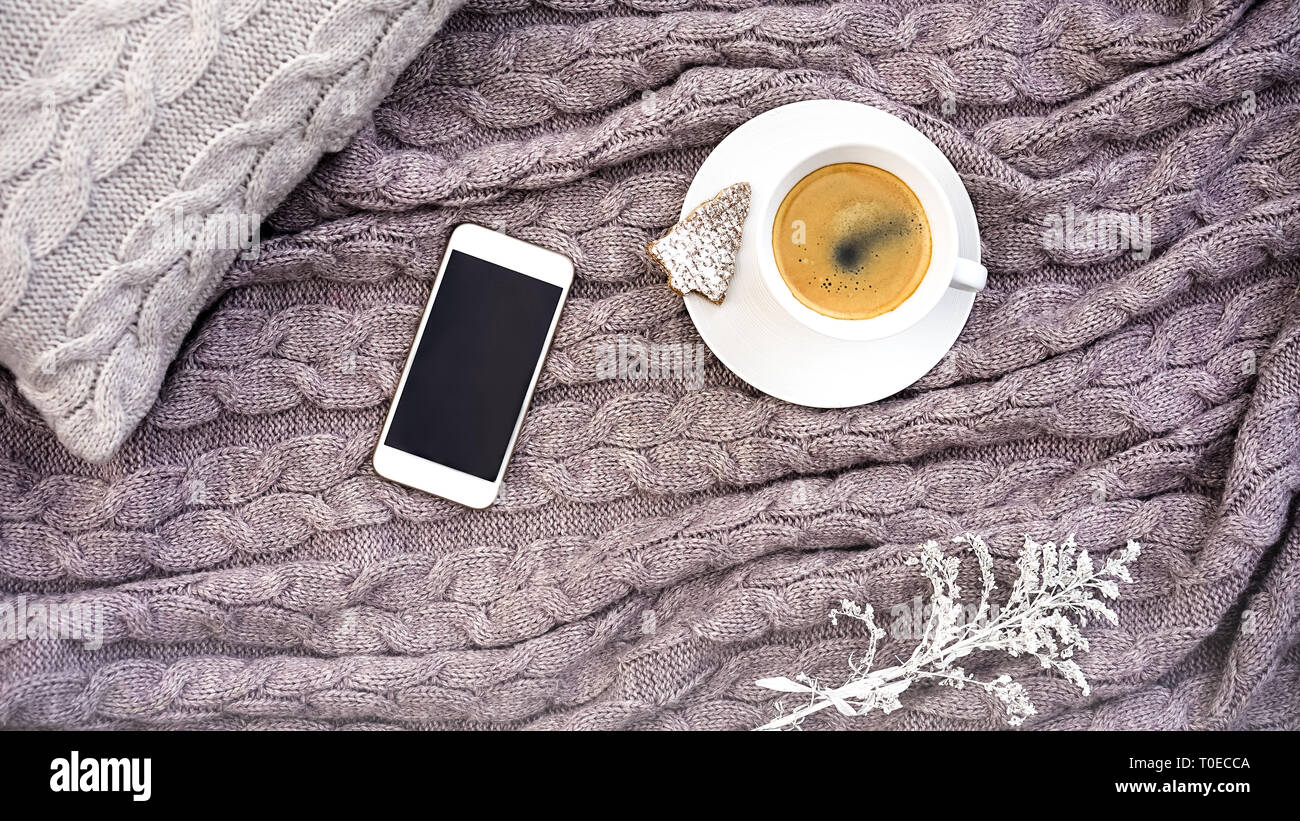 White tazza di caffè con i cookie nella forma di abete e telefono bianco fiore sul cuscino grigio e calda lana rosa grigio plaid. Tessile lavorato a maglia con motivo a treccia. Autunno Inverno concetto di comfort Foto Stock