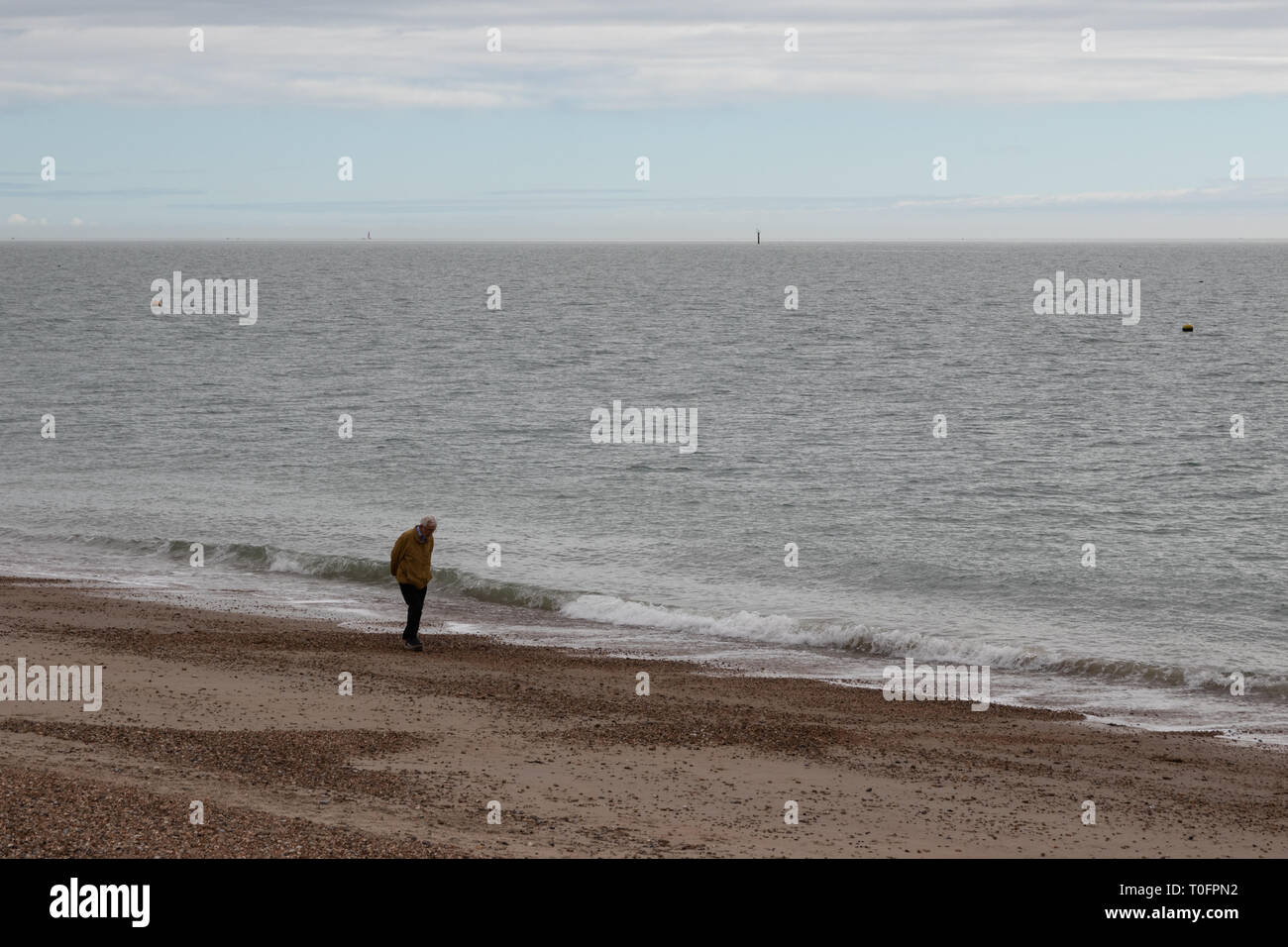Un anziano uomo a camminare da sola su una spiaggia di ciottoli con le mani dietro la schiena. Foto Stock