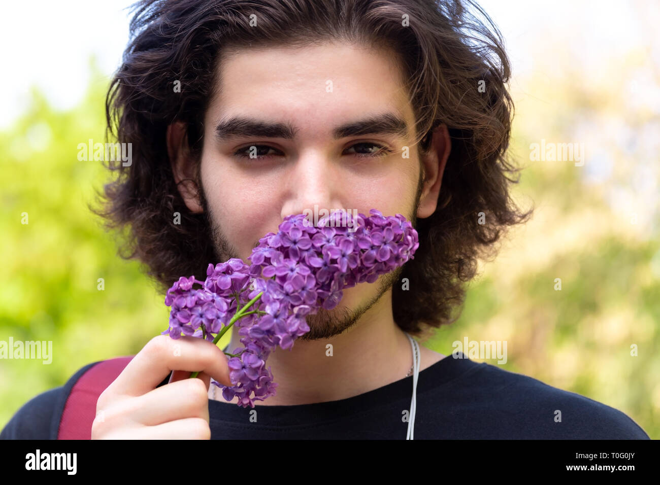 Bruna giovane ragazzo profumati fiori freschi sul ramo di lilla e guardando la fotocamera su sfondo sfocato Foto Stock