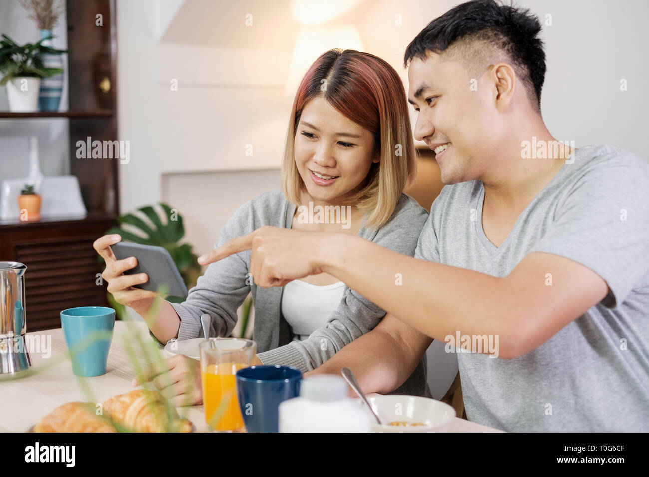 Felice coppia asiatica in pigiama seduti al tavolo in cucina e guardare video sul cellulare togerther a casa la mattina e avente la prima colazione di cereali. Foto Stock