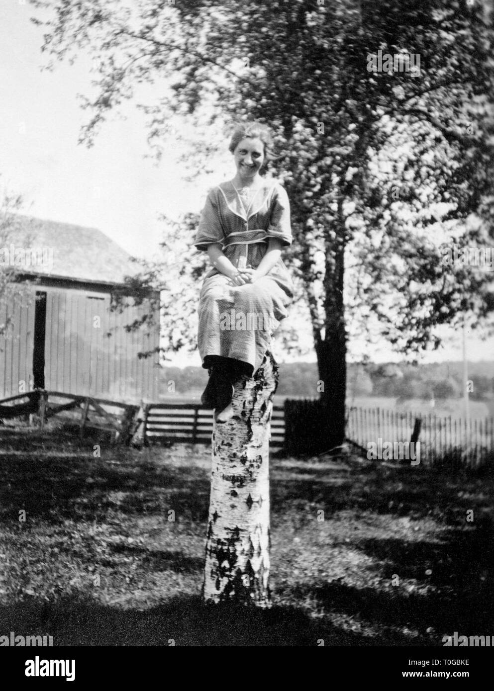 Una donna si siede in cima a un albero di betulla moncone nel cortile, ca. 1920. Foto Stock
