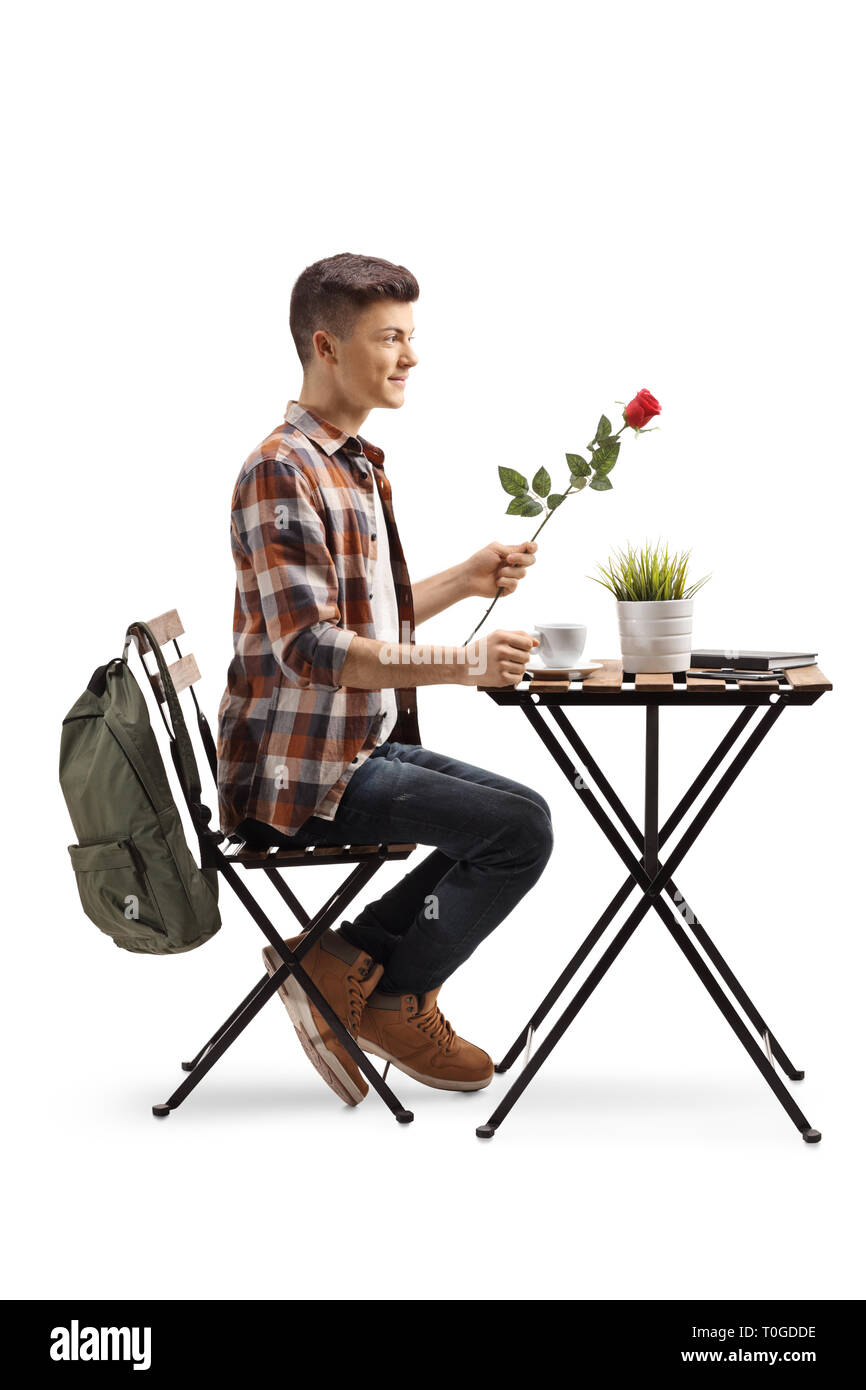 Lunghezza piena ripresa di profilo di un maschio di studente in possesso di una rosa rossa e seduti al tavolo con caffè isolato su sfondo bianco Foto Stock