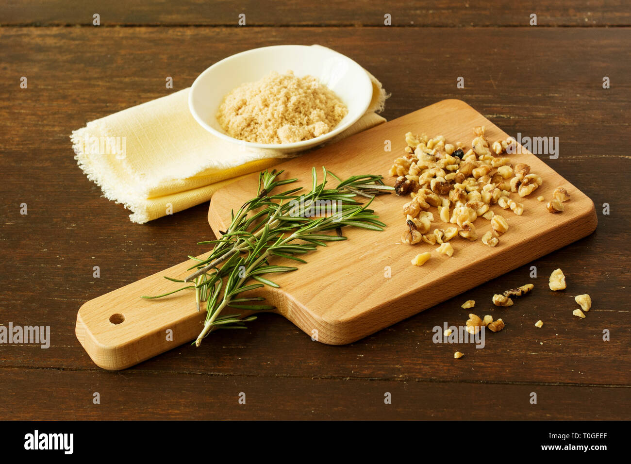 Noci tritate con alcuni rametti di rosmarino fresco su un tagliere di legno con una tazza di zucchero di canna su un distressed legno di ciliegio tavolo. Foto Stock