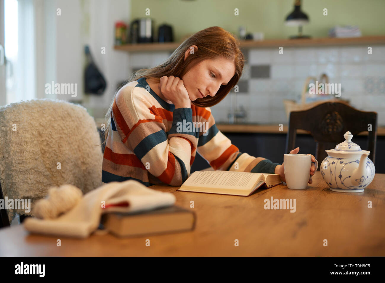 Bella donna bionda seduti ad un tavolo la lettura di un libro, sorridente, uncinetto gli utensili e i libri sul tavolo Foto Stock