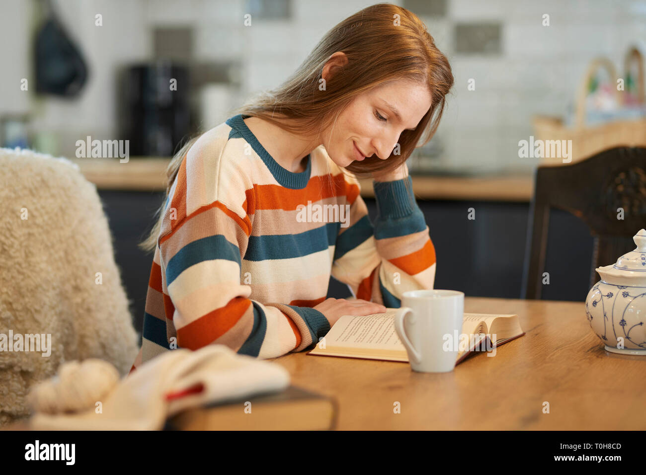 Bella donna bionda seduti ad un tavolo la lettura di un libro, sorridente, uncinetto gli utensili e i libri sul tavolo Foto Stock