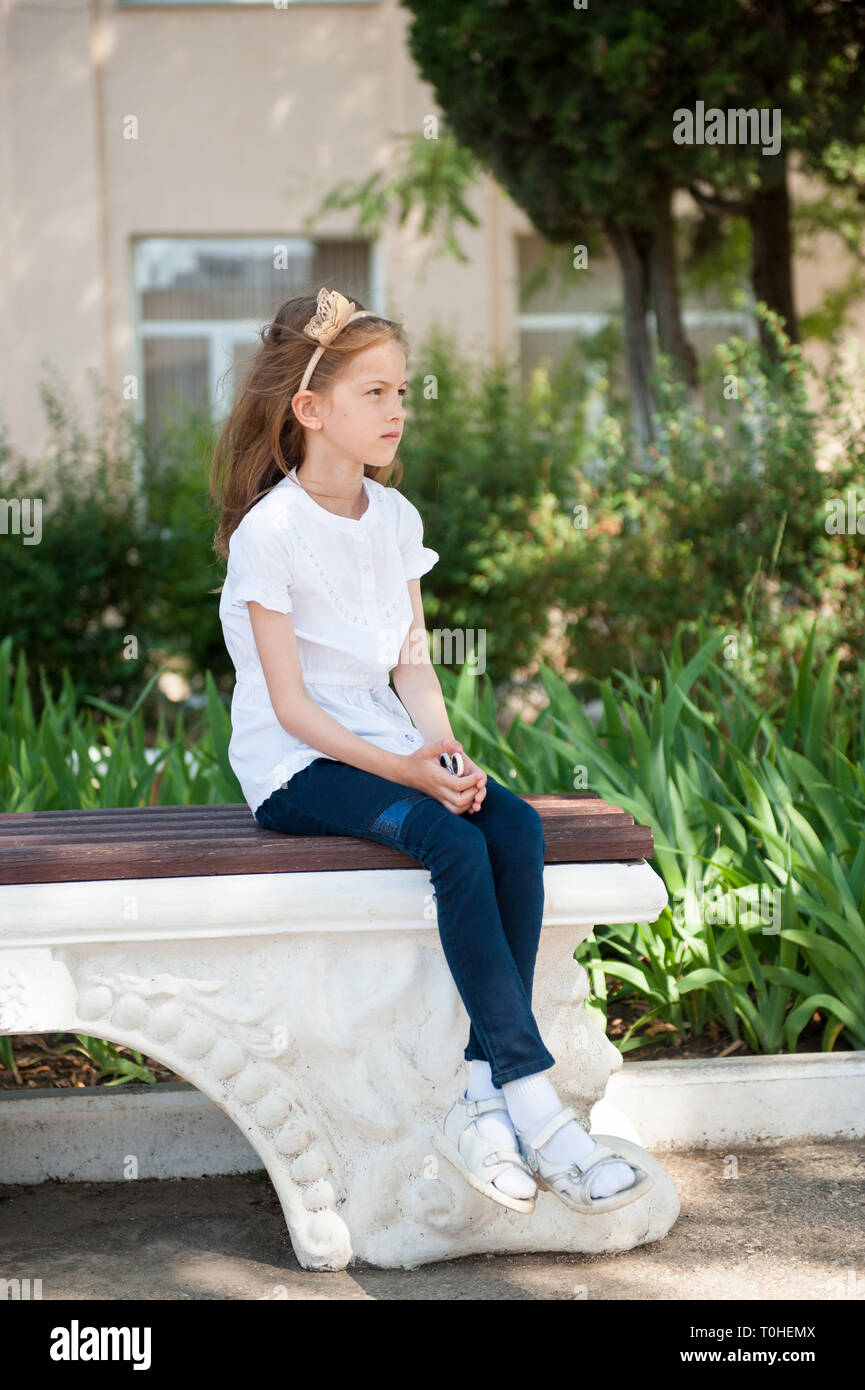 Una bella ragazza seduta sul banco vicino giardino verde in schoolyard Foto Stock