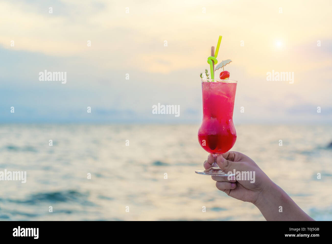 Mano azienda red cocktail drink in twilight sea & sky background. Estate, vacanze, viaggi e vacanze concetto. Foto Stock