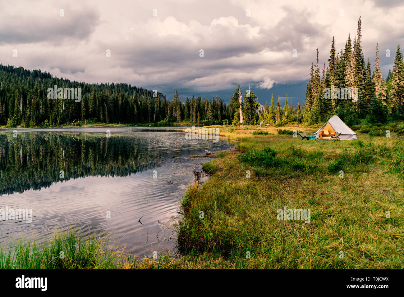 Campeggio con tenda illuminata accanto a incontaminato lago alpino nella foresta sempreverde. Foto Stock