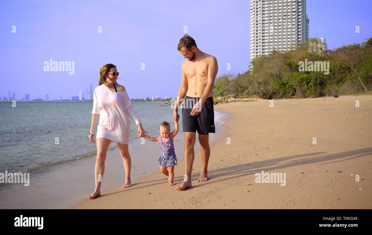 La famiglia felice in spiaggia, tenendo le mani, agitando una bambina intorno a. Mamma papà figlia a piedi sulla spiaggia. concetto di famiglia felice. 4k Foto Stock