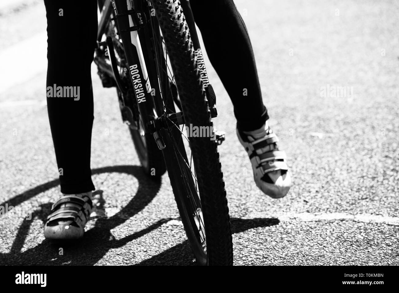 In bianco e nero a contrasto elevato e anteriore ruota di bicicletta con coppia di gambe su entrambi i lati della moto indossando scarpe da ciclismo, uno sollevato e uno sul terreno Foto Stock
