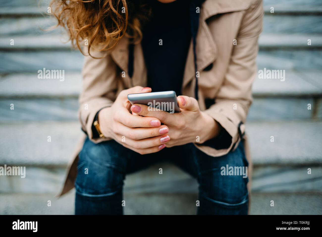 Close-up giovane donna che indossa il beige trench coat e jeans blu seduti sulla pietra grigia scale utilizzando smart phone. La femmina mani tenendo il dispositivo mobile e Foto Stock