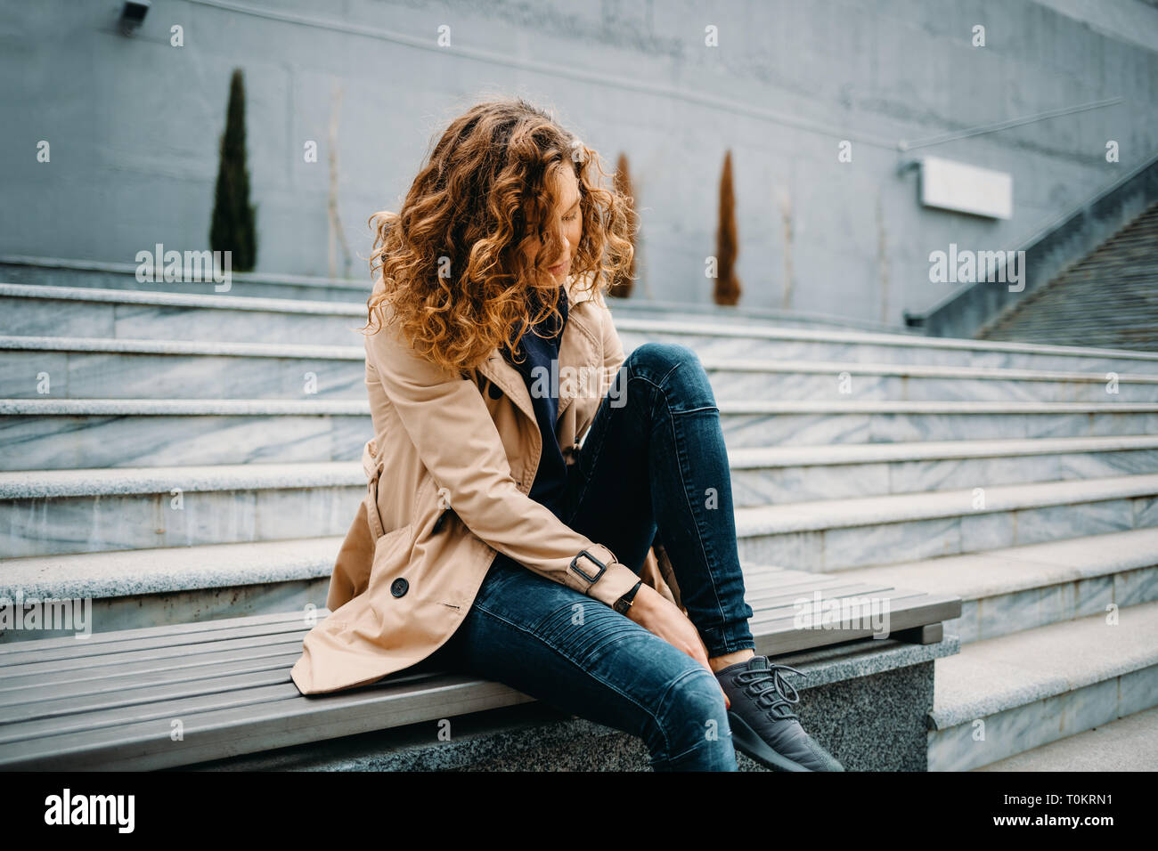 Giovane donna che indossa il beige trench coat e jeans blu seduti sulla  pietra grigia scale adegua la sua sneakers Foto stock - Alamy