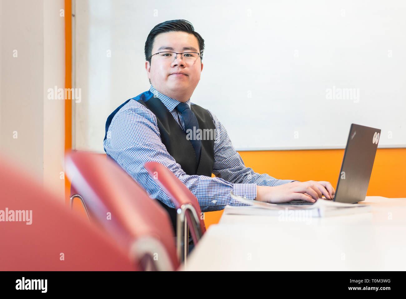 Un internazionale asiatico maschio giovane studente si siede in una classe a un banco di lavoro o di studio sul suo computer portatile in una moderna classe. Foto Stock