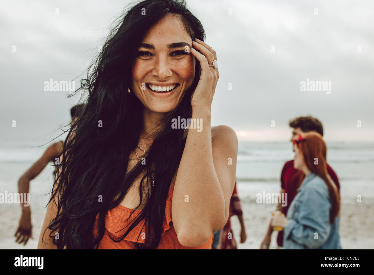 Attraente giovane donna sulla spiaggia sorridente. Bella femmina caucasica in piedi presso la spiaggia con un gruppo di amici in background. Foto Stock