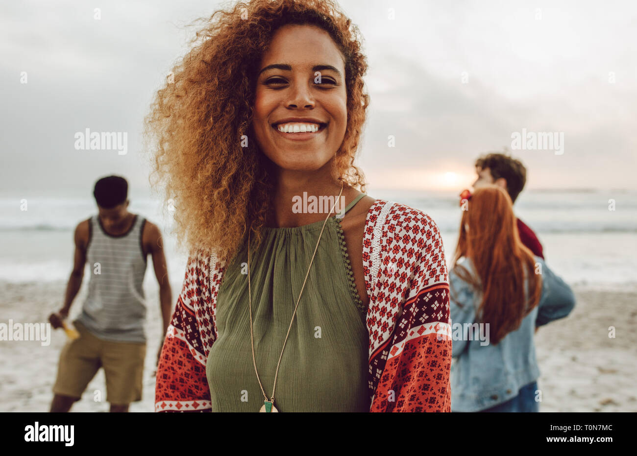 Attraente donna africana sulla spiaggia di sorridere alla telecamera. Piuttosto giovane donna in piedi presso la spiaggia con un gruppo di amici in background. Foto Stock