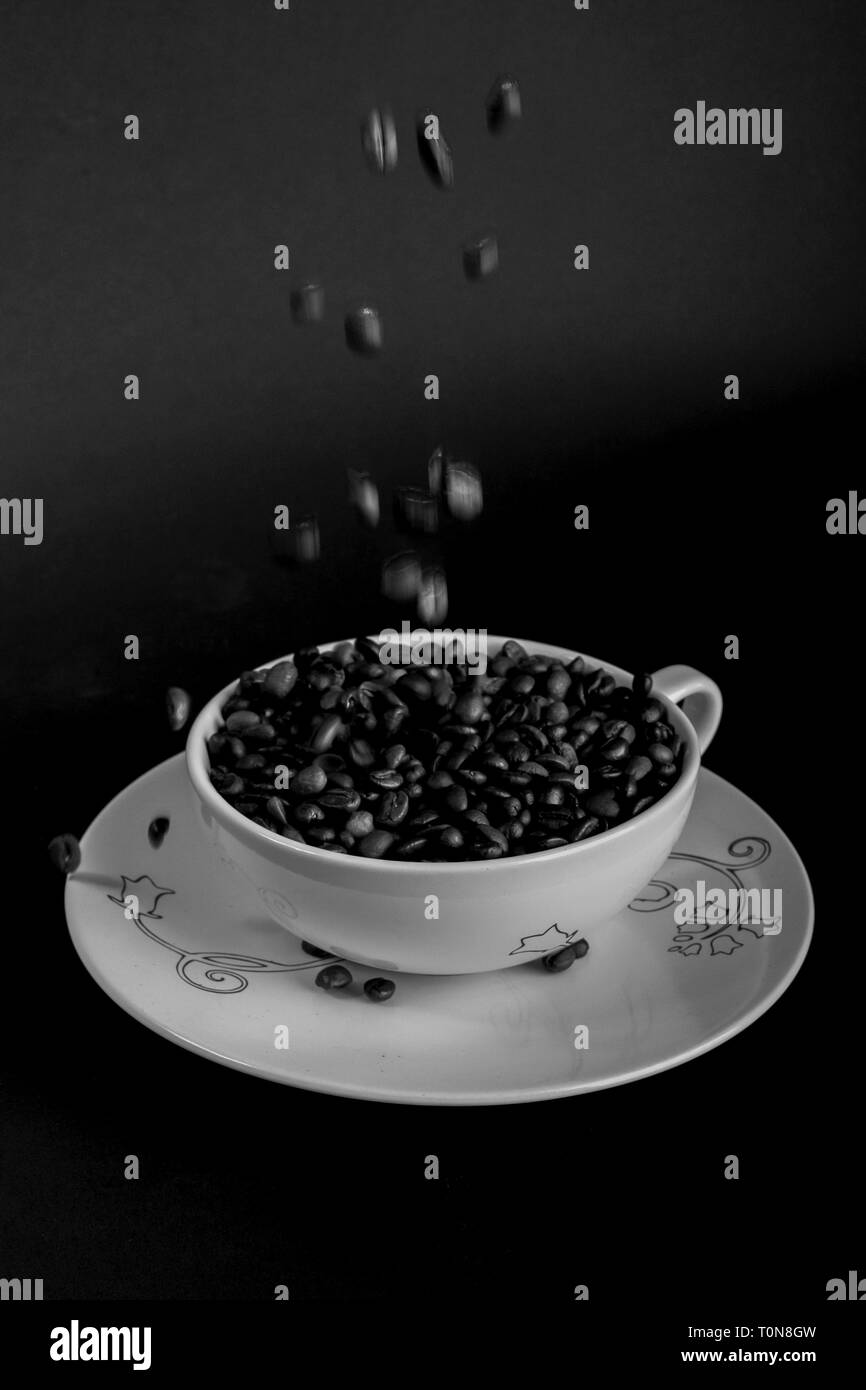 Concetto che mostra i chicchi di caffè che viene versato in un bianco tazza da caffè con piattino su sfondo nero Foto Stock