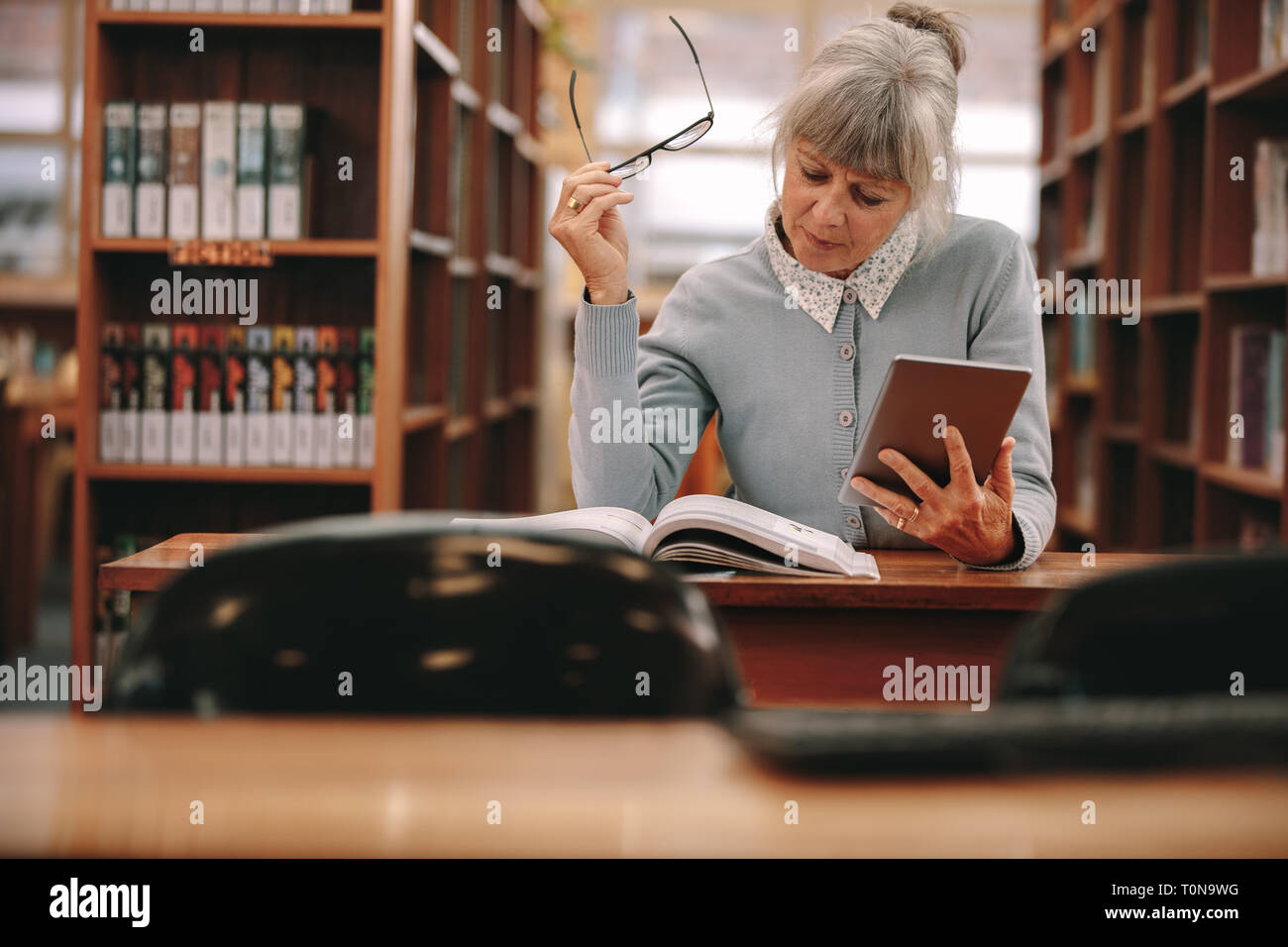 Senior donna utilizzando un tablet pc per riferimento durante la lettura di un libro in biblioteca. Donna seduta in una libreria la lettura di un libro. Foto Stock