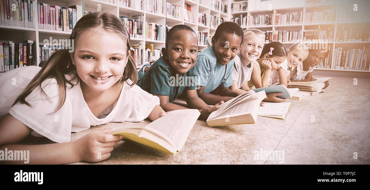 Scuola sorridente kids giacente sul piano la lettura di libro in biblioteca Foto Stock