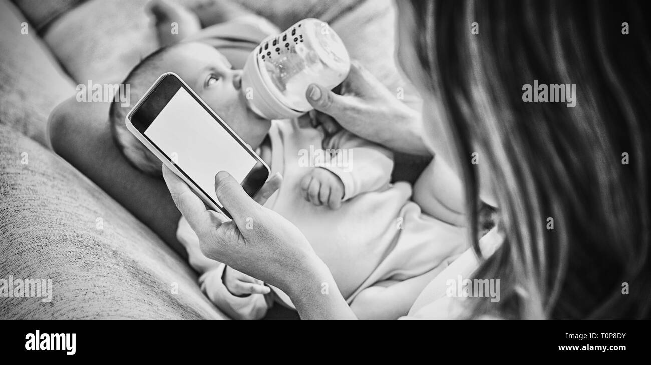 Madre utilizzando il telefono cellulare mentre si alimenta il suo bambino con bottiglia di latte Foto Stock