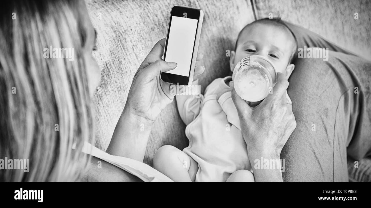 Madre utilizzando il telefono cellulare mentre si alimenta il suo bambino con bottiglia di latte Foto Stock