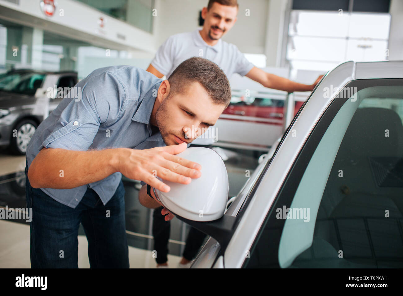 Attento ai dettagli cliente si appoggia sulla vettura bianca e tocca a lei. Giovane uomo guarda nel mirror. Egli è grave e concentrata. Il venditore si erge dietro di lui un Foto Stock
