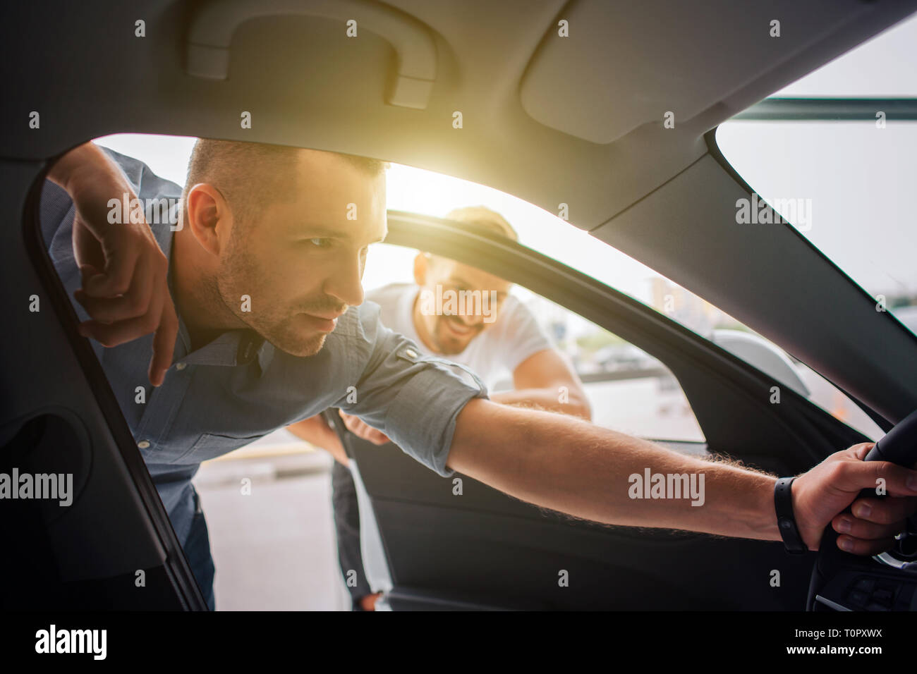 Bello e sicuro il cliente si alza e si appoggia alle auto. Egli tiene il volante con una sola mano. Il ragazzo è attento ai dettagli. Manager sta dietro di lui Foto Stock