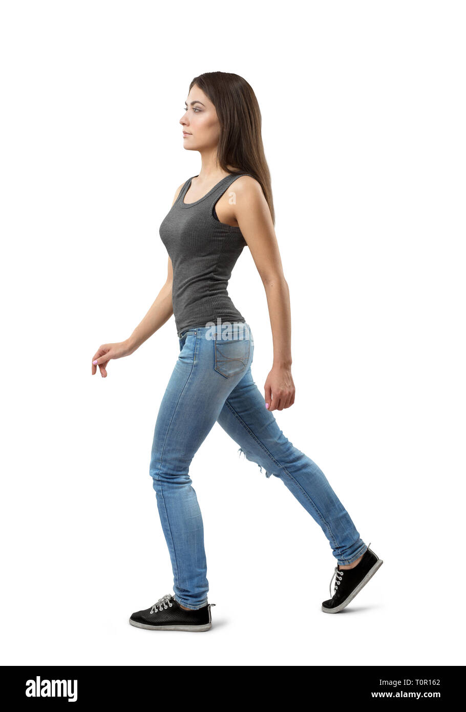 Vista laterale della giovane donna in grigio top senza maniche e jeans blu, con lunghi nocciola scuro capelli, a camminare in avanti su sfondo bianco. Foto Stock