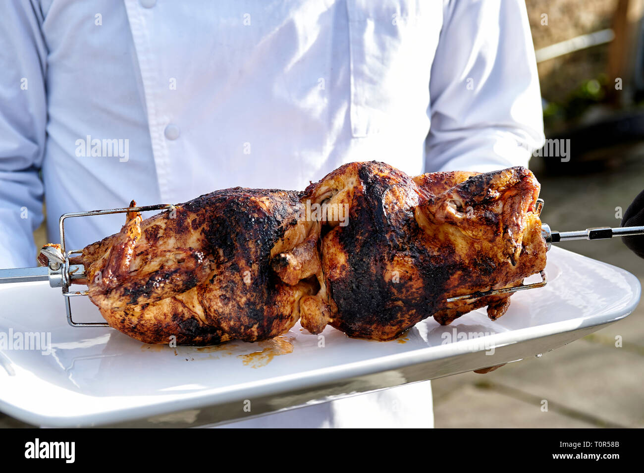 Grillen mit Gefluegel: Ein Koch serviert zwei Grillhaehnchen die noch auf dem Grillspiess sind. Foto Stock