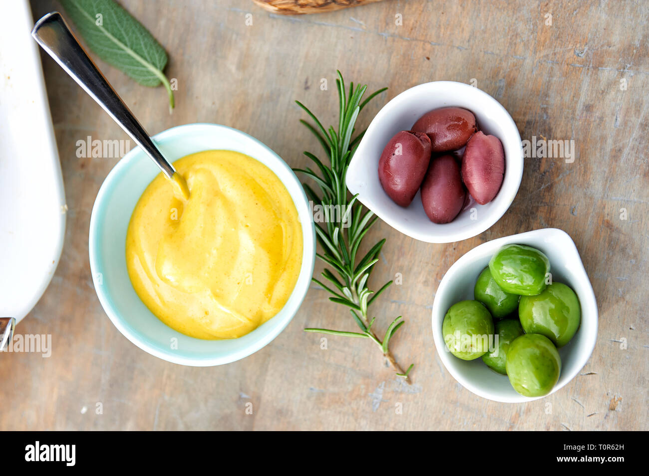 3 Schaelchen mit gruenen und schwarzen Oliven, sowie Majonnaise stehen auf einem Tisch. Deko: ein Salbeiblatt und ein Rosmarienzweig und Loeffel Foto Stock