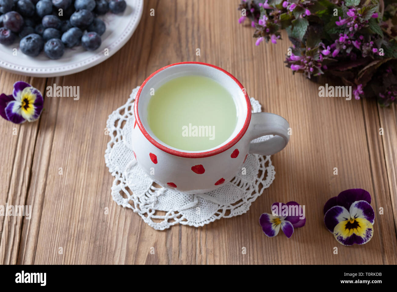 Il siero di latte fresco in una tazza con mirtilli, pansy fiori viola e dead-ortiche in background Foto Stock