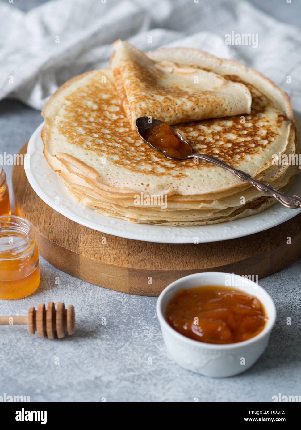 Pila russo in casa il lievito Pancake sulla piastra bianca, legno cucchiaio di miele, miele in vasetti e confettura di albicocca nella ciotola sul tavolo. Tradizionale frittelle di frumento per Foto Stock