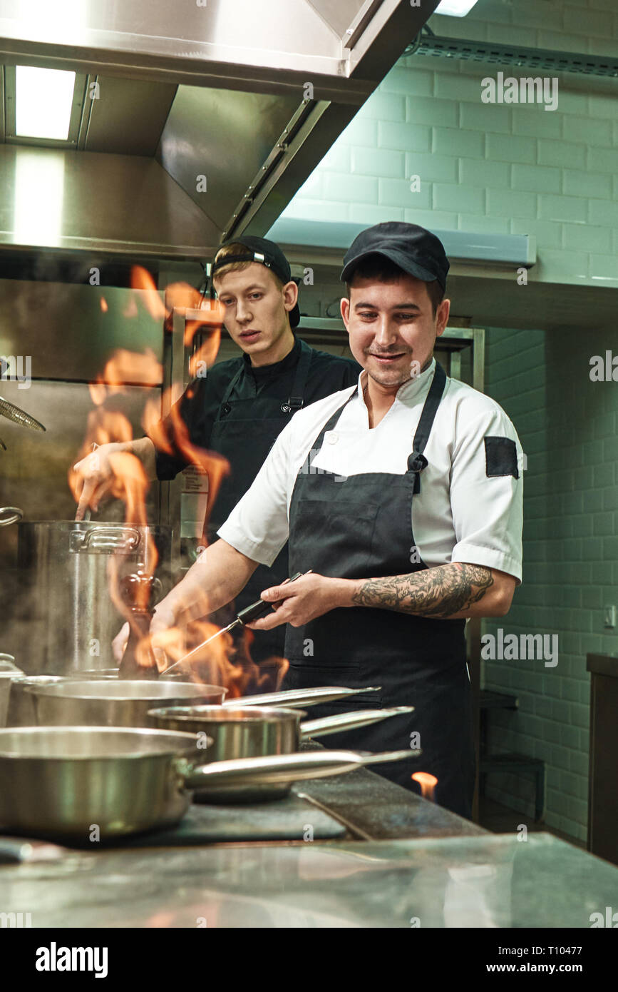 Lavorando con il fuoco. Due giovani e cuochi positivo per imparare a fare un flambe sul cibo del ristorante cucina. Scuola di cucina. Il lavoro di squadra. Foto Stock