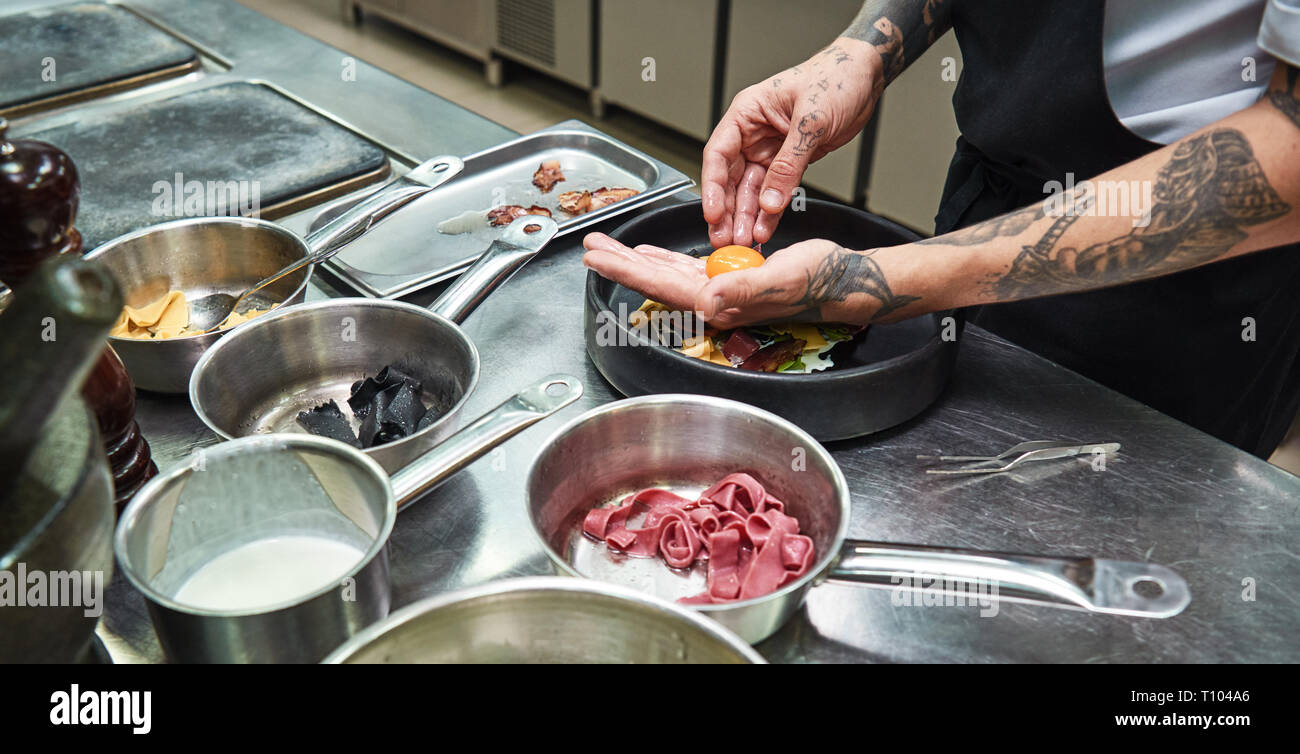 Ingrediente speciale. Immagine ritagliata dello chef mani con parecchi tatuaggi aggiungere un tuorlo alla pasta italiana mentre in piedi in un ristorante di cucina Foto Stock