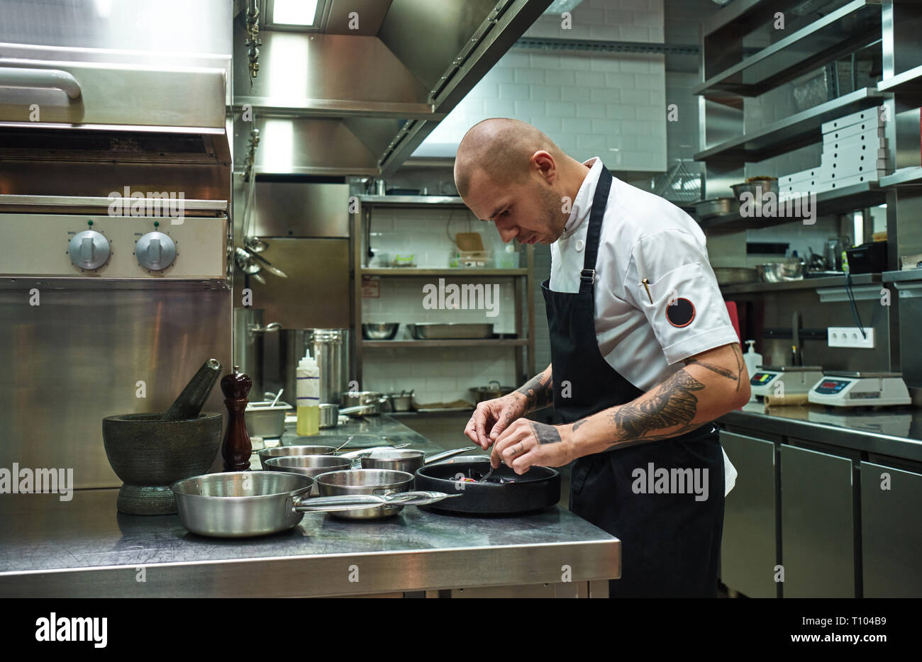 Decorazione di prodotti alimentari. Vista laterale del fiducioso chef maschio con parecchi tatuaggi sulle sue braccia guarnendo il suo piatto in un ristorante di cucina. Concetto di cucina a vista Foto Stock