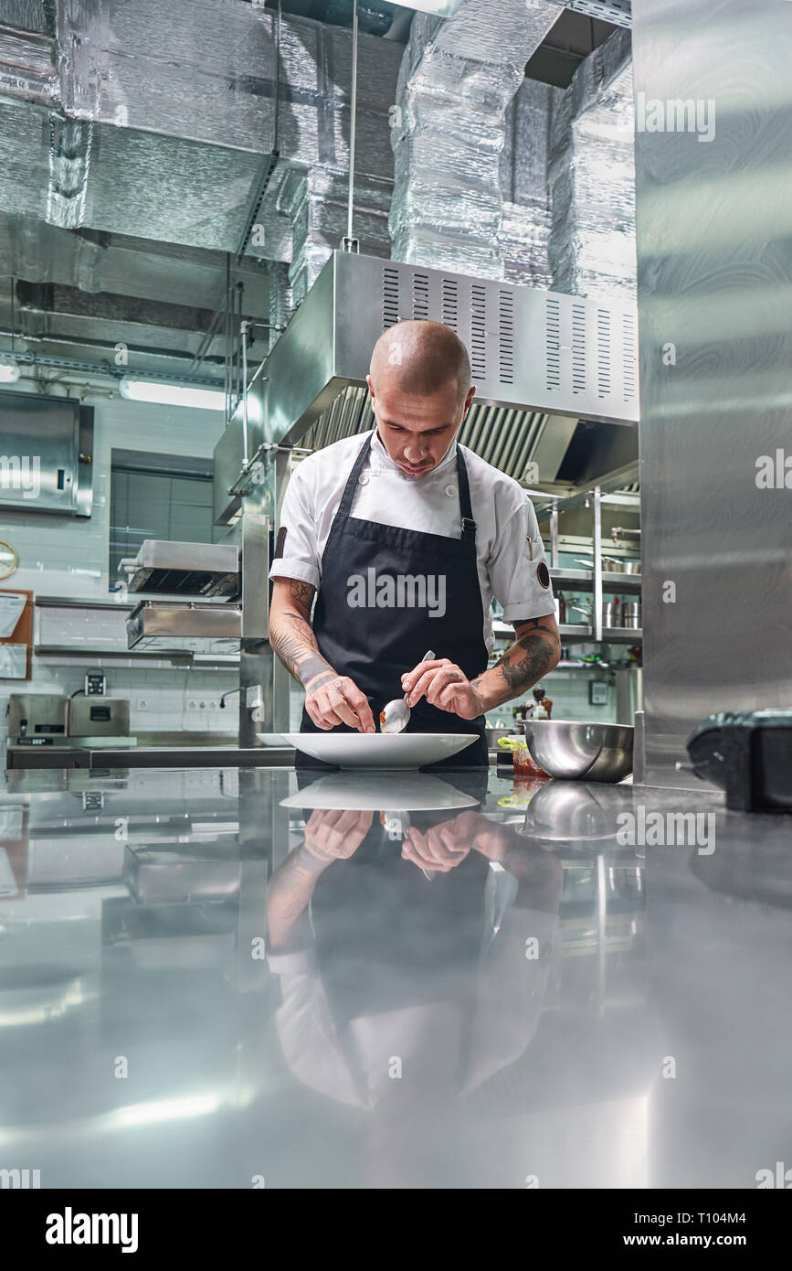 Lavorando in un ristorante. Ritratto verticale del maschio professionale chef con tatuaggi sulle sue braccia guarnendo il suo piatto della piastra bianca mentre in piedi in una cucina Foto Stock