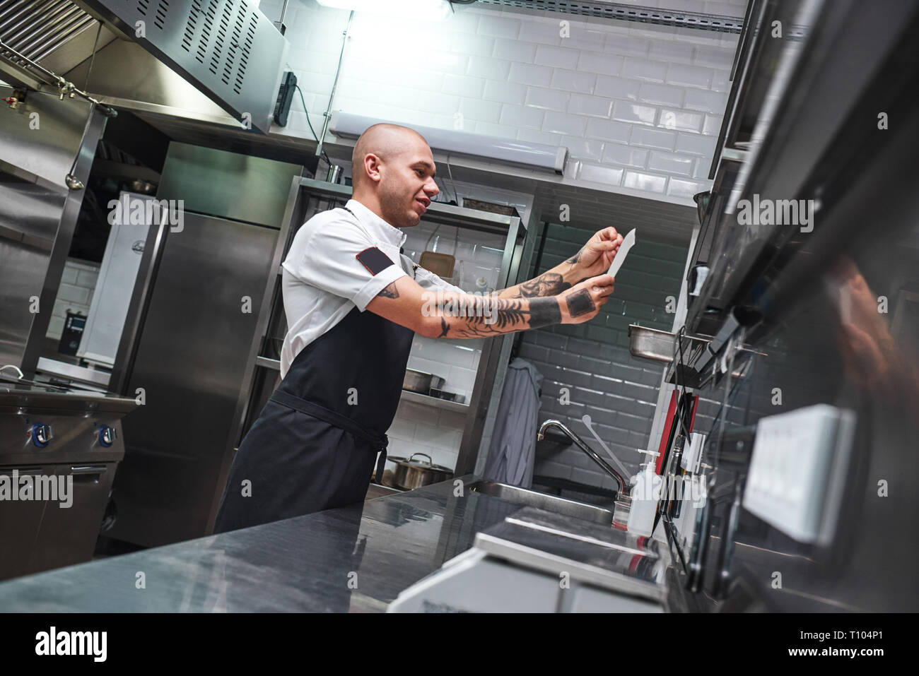Qual è il prossimo... Bello calvo chef maschio con tatuaggi sulle sue braccia guardando lista ordine in un ristorante di cucina. Concetto di cucina a vista Foto Stock