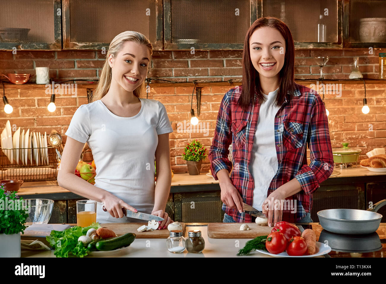 Ritratto di due donne attraenti, divertendosi, durante la preparazione di insalata di verdure. Essi sono pienamente coinvolti nel processo. Essi stanno guardando la telecamera e sorridere mentre per affettare i funghi. Vista frontale Foto Stock
