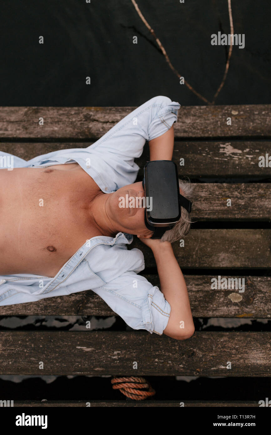 Senior uomo disteso sul pontile di un lago indossando occhiali VR Foto Stock