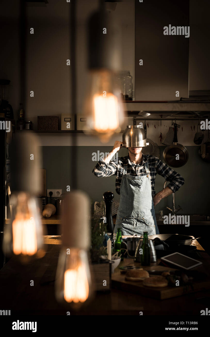 Uomo con grembiule in piedi in cucina, indossando scolapasta come casco Foto Stock