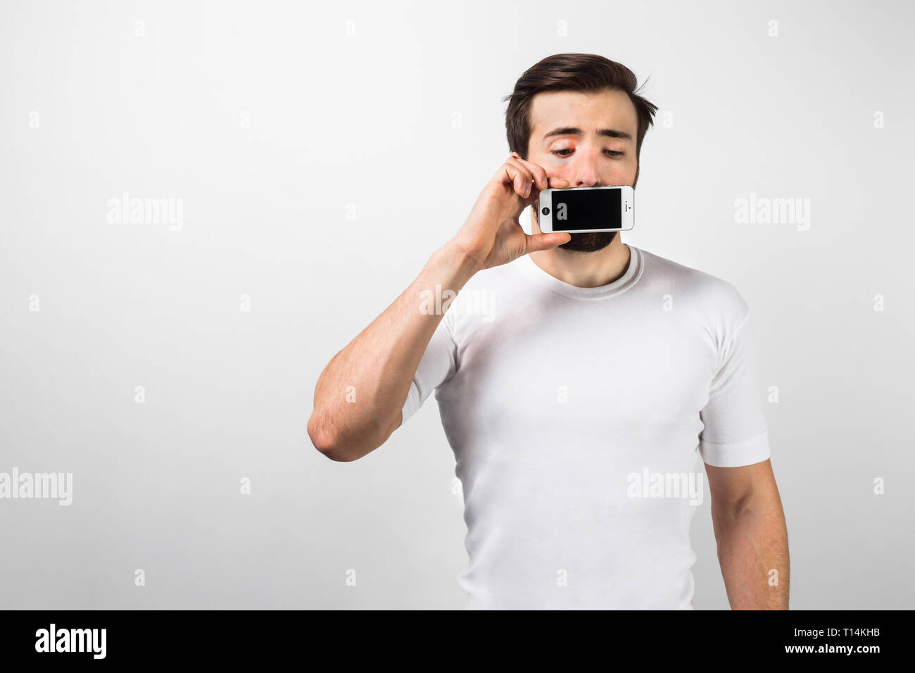 Un interessante quadro di un ragazzo in piedi vicino al grande muro bianco e chiudendo la bocca con un telefono nella sua mano destra. Egli guarda distured e preoccupato. isol Foto Stock