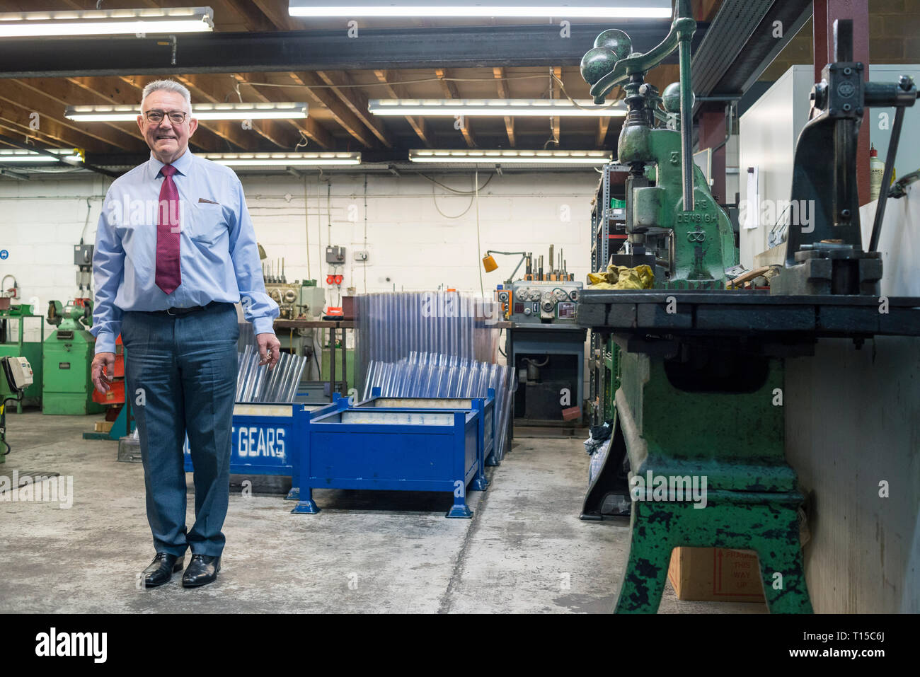 Un simpatico anziano signore / titolare di azienda in piedi e sul piano della fabbrica del suo tradizionale azienda manifatturiera circondato da strumenti Foto Stock