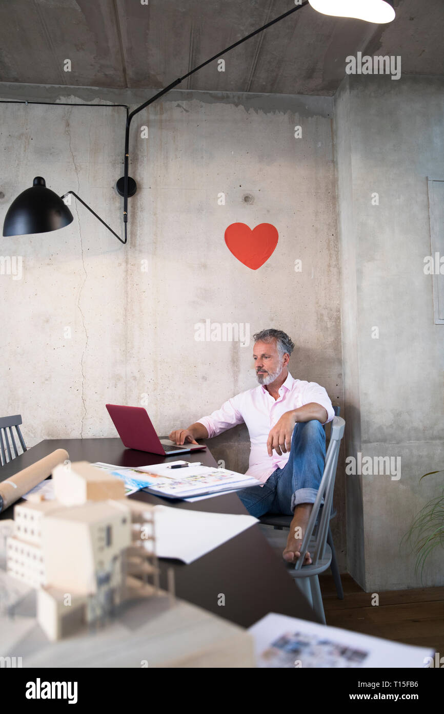 Imprenditore in un loft utilizzando laptop con documenti e modello architettonico sulla tavola e il cuore sulla parete Foto Stock